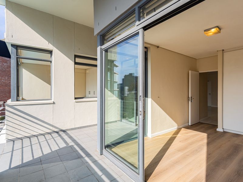 A bright, empty balcony with tiled flooring and metal railings, seamlessly connected to an interior living space through a large sliding glass door.