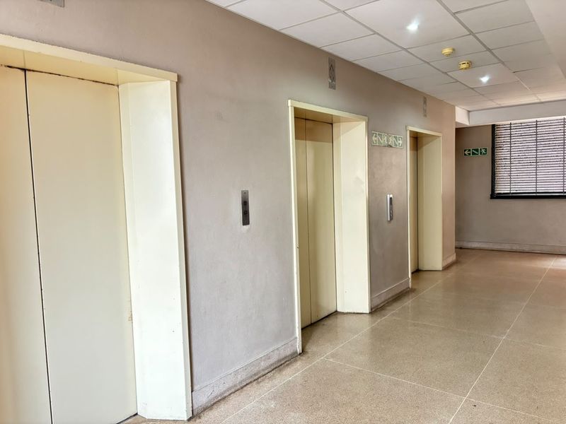 A functional apartment building hallway featuring multiple elevator access points, tiled flooring, and a suspended ceiling with recessed lighting.