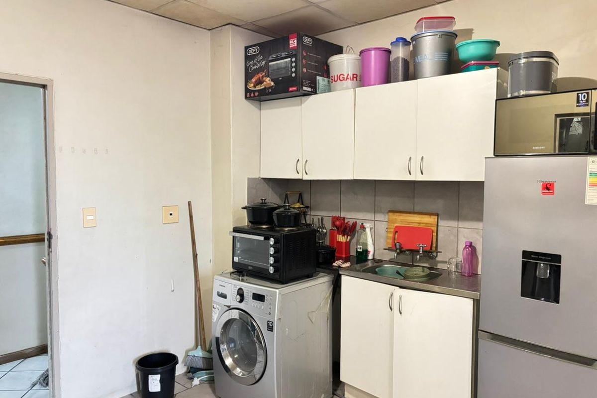 A compact kitchen area featuring white built-in cabinetry, a stainless steel sink, and tiled backsplash, requiring cosmetic updates to improve its overall condition.