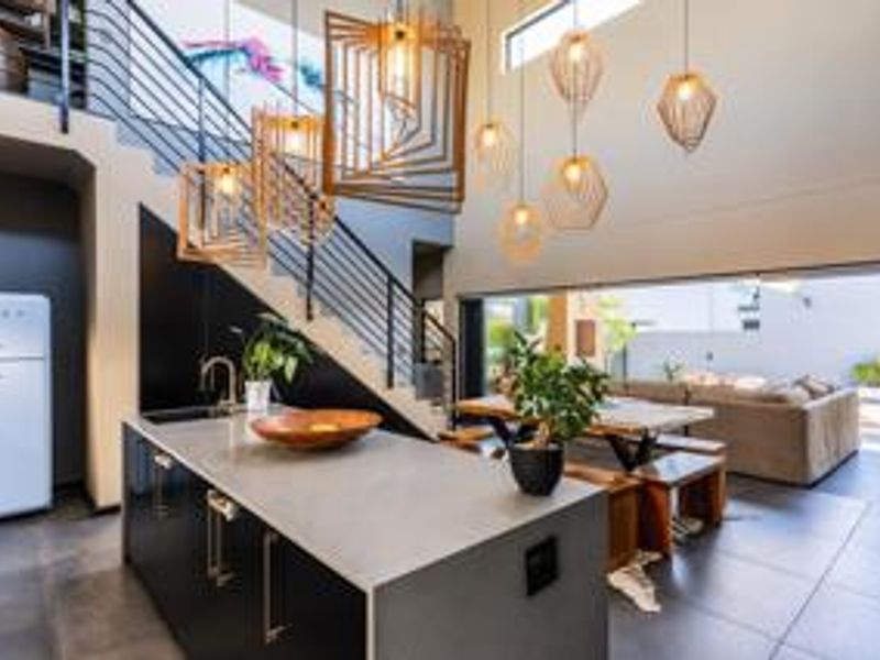 Modern open-plan living area featuring a sleek kitchen island, dramatic double-volume ceiling with pendant lighting, and seamless flow to the outdoor patio.