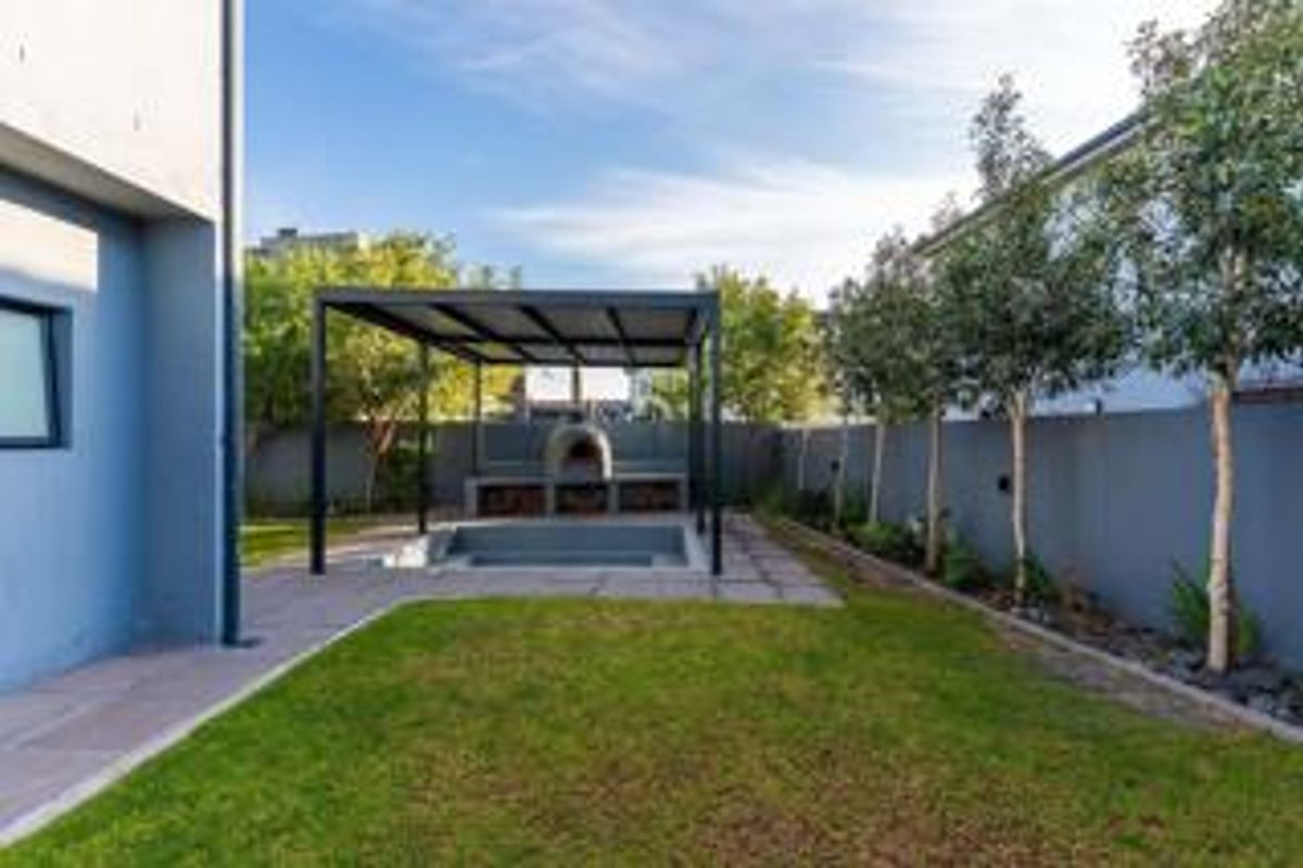 A well-maintained rear garden featuring a paved patio area with a built-in pizza oven and braai, enclosed by a boundary wall.