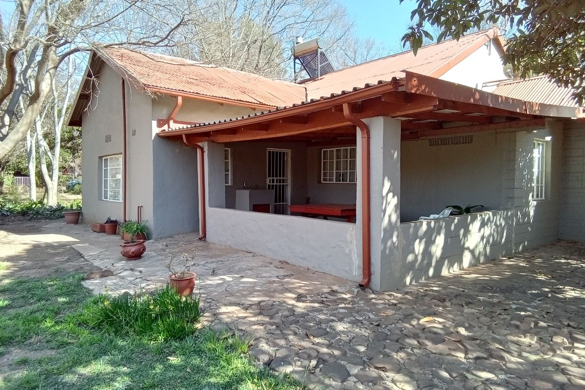 Rear view of the farmhouse featuring a covered patio with a built-in braai, stone paved area, and a corrugated metal roof with solar heating.