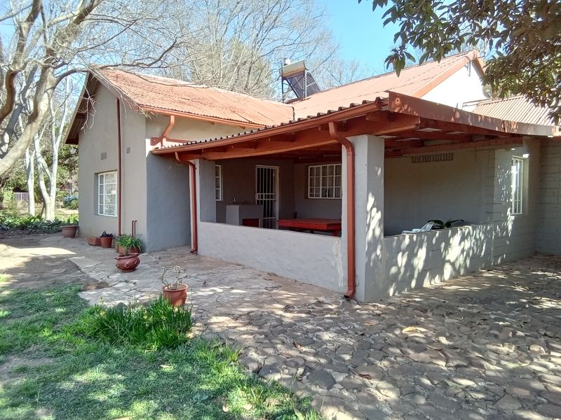 Rear view of the farmhouse featuring a covered patio with a built-in braai, stone paved area, and a corrugated metal roof with solar heating.
