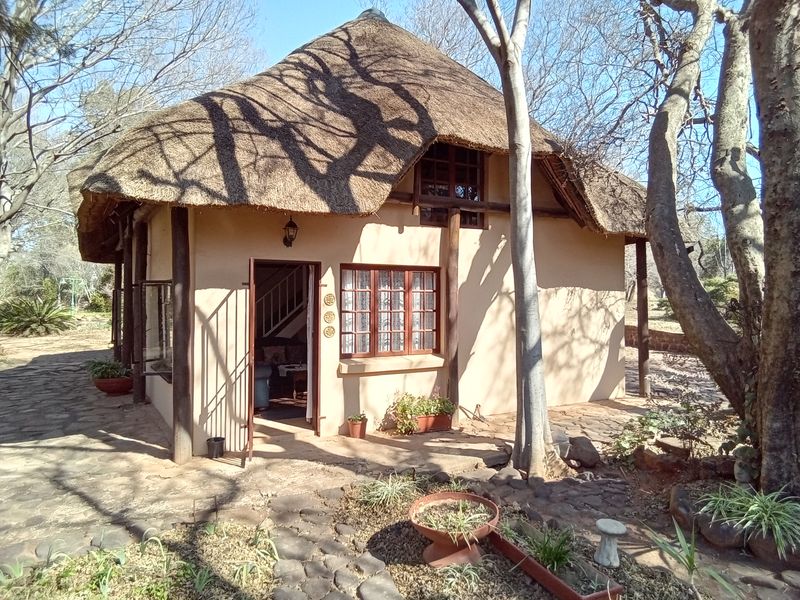 A charming cottage with a traditional thatched roof, timber structural elements, and stone paving, set within a natural, tree-lined farm environment.