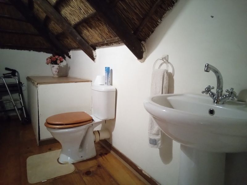 A rustic bathroom featuring a pedestal basin, toilet, and exposed timber rafters under a traditional thatched roof with timber flooring.