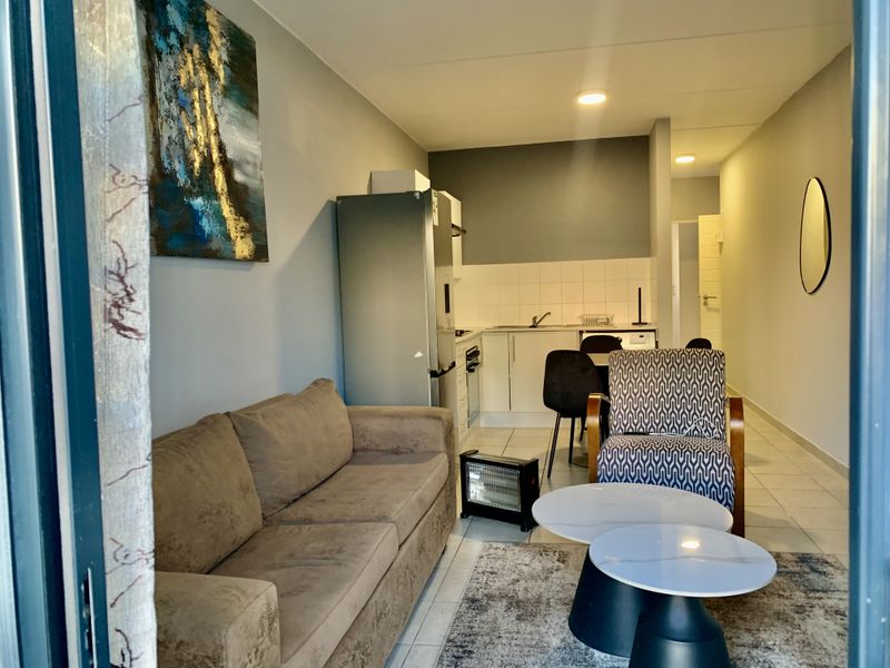 An open-plan living and kitchen area featuring tiled flooring, neutral wall tones, and a functional kitchenette with built-in cabinetry and appliances.