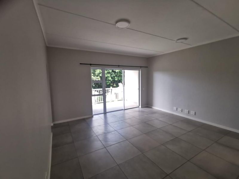 An empty, neutral-toned living room featuring large floor tiles and a sliding glass door providing access to an outdoor balcony area.
