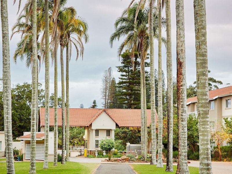 A well-maintained apartment complex entrance featuring a paved driveway, mature palm trees, and a secure gated access point with tiled roof structures.