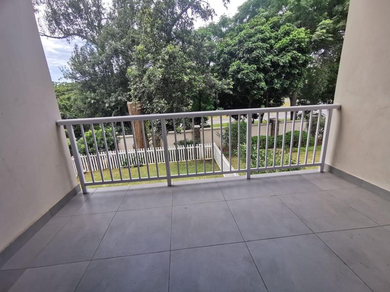 A clean, covered balcony featuring modern grey floor tiles and a white metal balustrade, overlooking a lush green garden area.