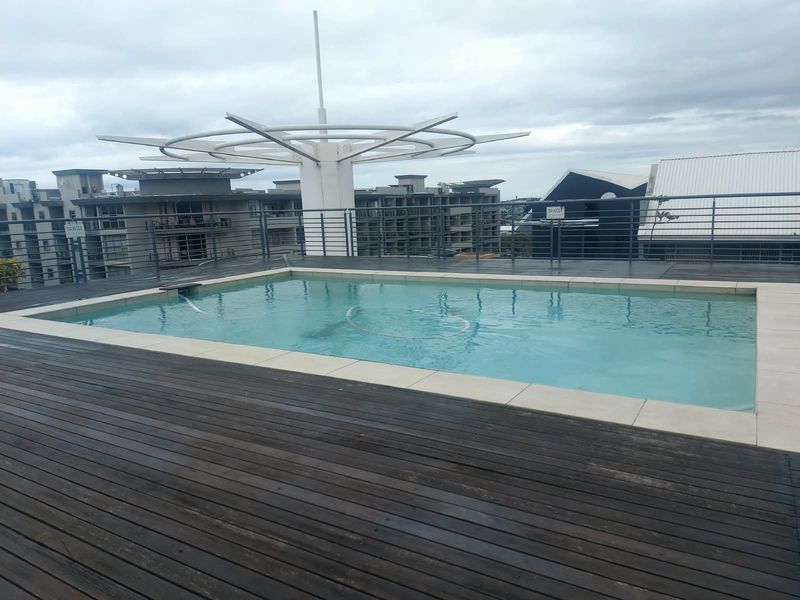 Rooftop swimming pool area featuring timber decking, metal safety railings, and a central architectural shade structure.