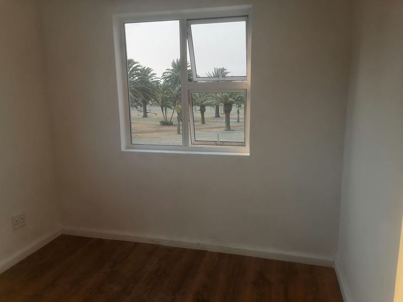 An empty bedroom featuring laminate flooring, neutral walls, and an aluminium window with views of palm trees.