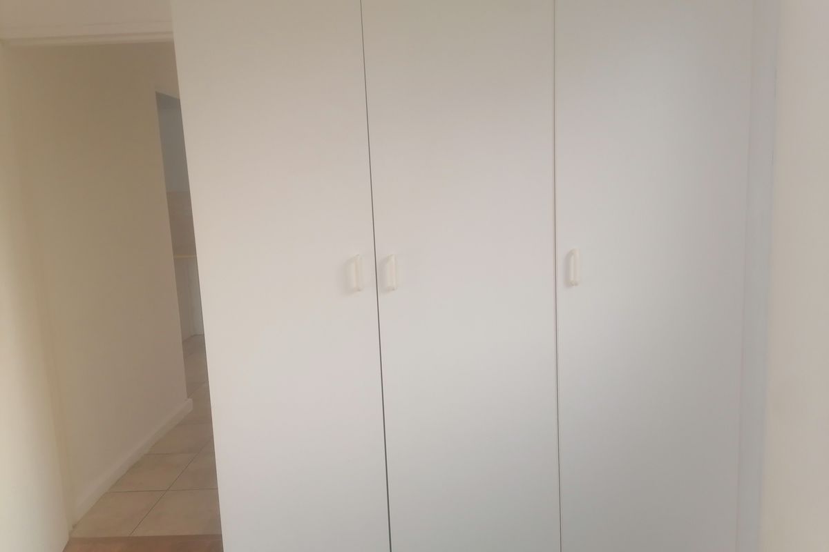 A bedroom interior featuring a white built-in wardrobe with simple handles, showing a view into an adjacent tiled hallway.