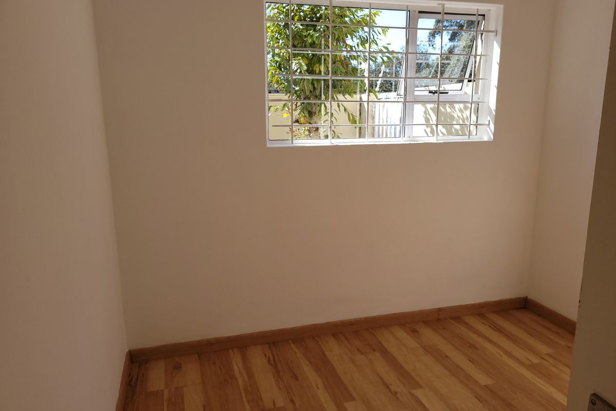 A bright, empty bedroom featuring laminate wood flooring and a window fitted with security bars.