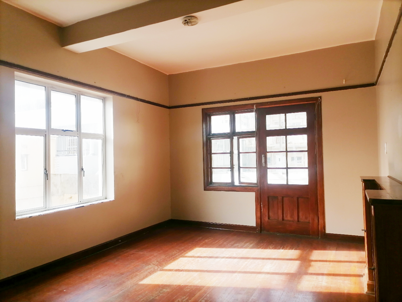 An empty living room featuring original hardwood flooring, timber-framed windows, and a classic timber door, offering a blank canvas for renovation.