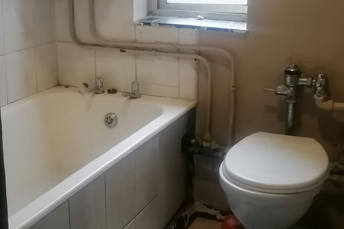 A dated bathroom featuring a bathtub and toilet with exposed plumbing and visible wear to the wall finishes and tiling.