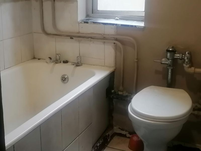 A dated bathroom featuring a bathtub and toilet with exposed plumbing and visible wear to the wall finishes and tiling.