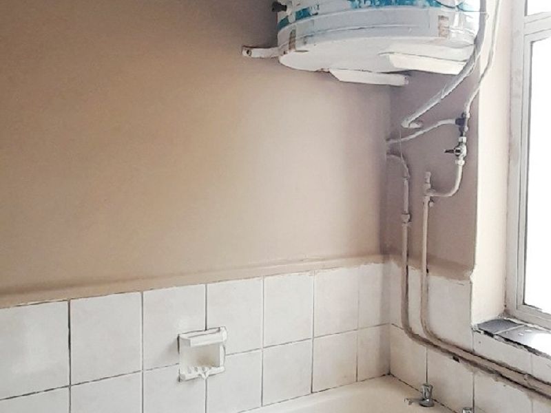 A dated bathroom featuring a bathtub and a wall-mounted electric geyser with visible plumbing, requiring modernization and maintenance of finishes.