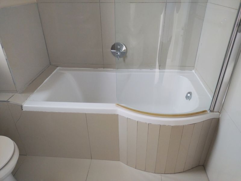 A functional bathroom featuring a built-in bathtub with a glass shower screen, neutral wall and floor tiling, and a wall-mounted mixer tap.