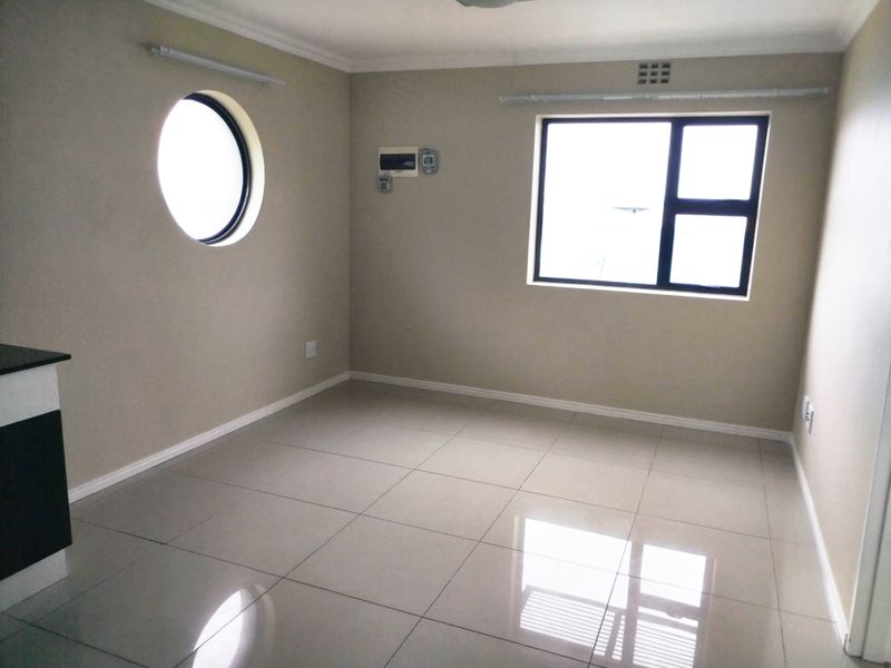 A bright, empty living area featuring polished porcelain floor tiles, a unique circular window, and a standard rectangular window with neutral wall finishes.
