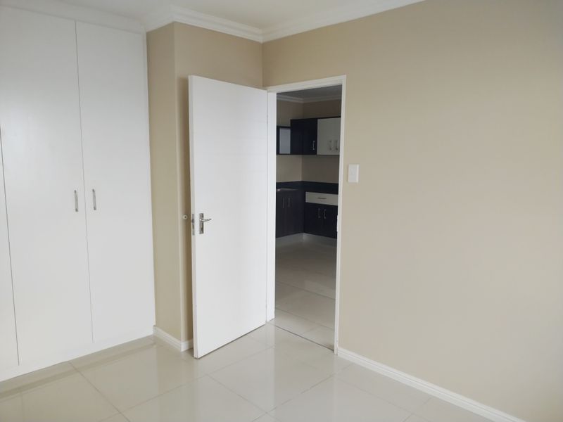 A clean, empty bedroom featuring neutral walls, tiled flooring, and built-in cupboards, with a view into an adjacent kitchen area.
