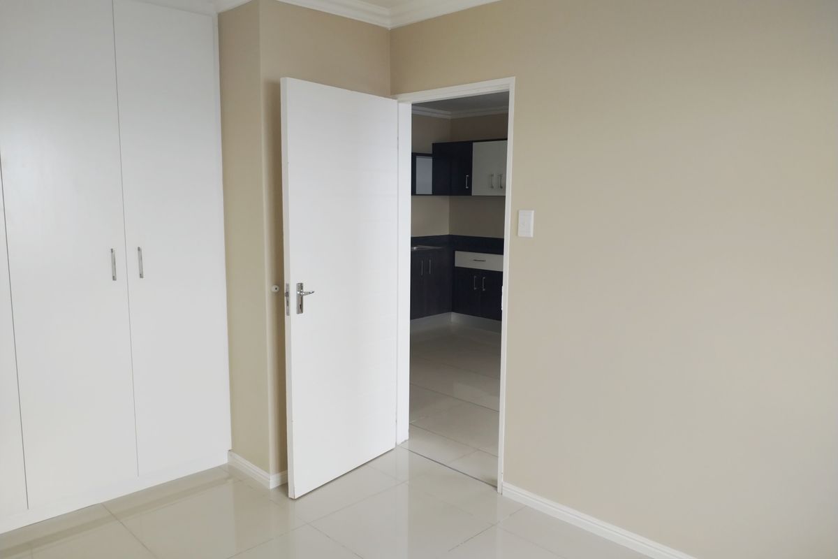 A clean, empty bedroom featuring neutral walls, tiled flooring, and built-in cupboards, with a view into an adjacent kitchen area.