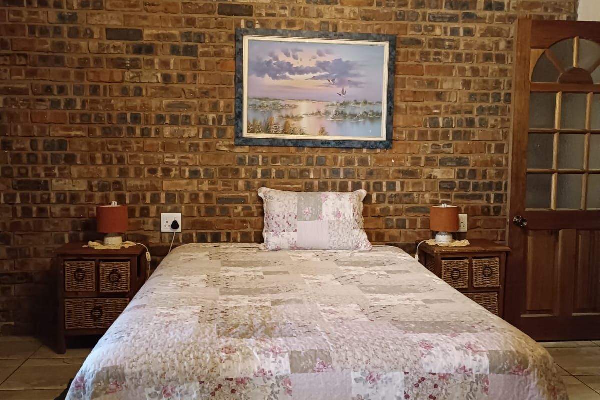 A rustic bedroom featuring an exposed face-brick feature wall, tiled flooring, and a wooden ceiling with a traditional chandelier.
