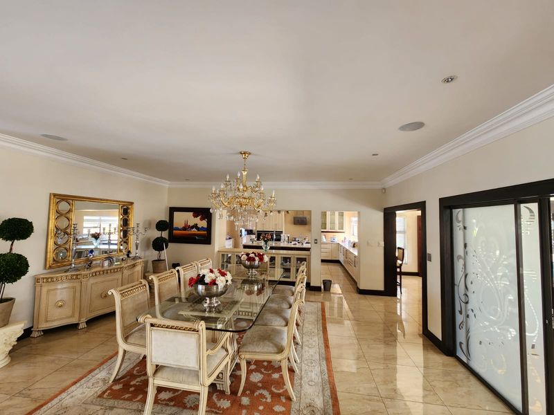 Spacious formal dining room featuring elegant crown molding, polished tile flooring, a grand crystal chandelier, and seamless flow into the adjacent kitchen area.