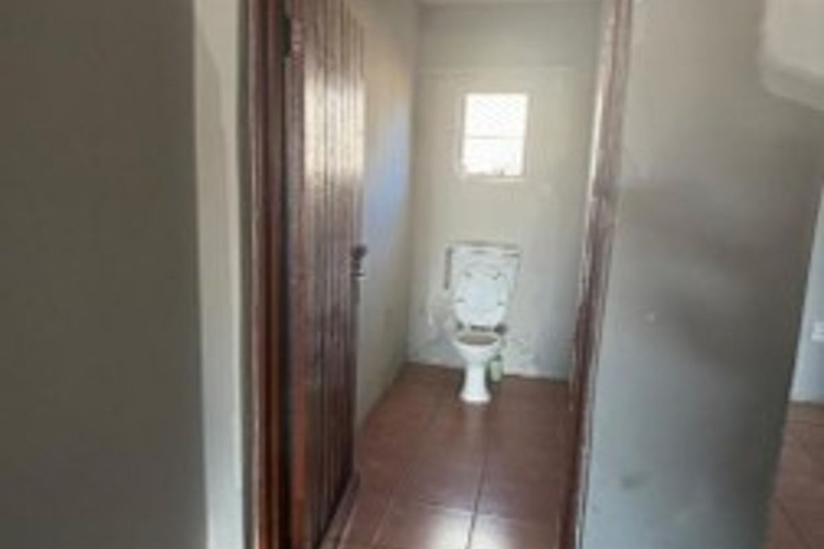 A small, functional bathroom featuring a toilet, tiled flooring, and unfinished wall surfaces, viewed from an adjacent hallway.