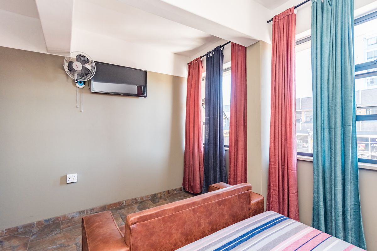 A bedroom featuring tiled flooring, large windows with curtains, and a wall-mounted fan, providing a functional and well-lit living space.