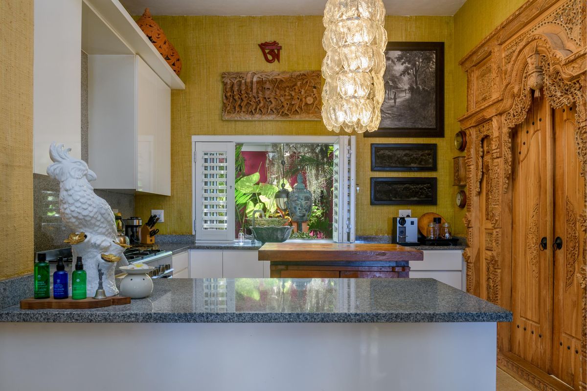 A well-appointed kitchen featuring granite countertops, white cabinetry, and a unique carved wooden door frame, overlooking a lush garden through a window with shutters.