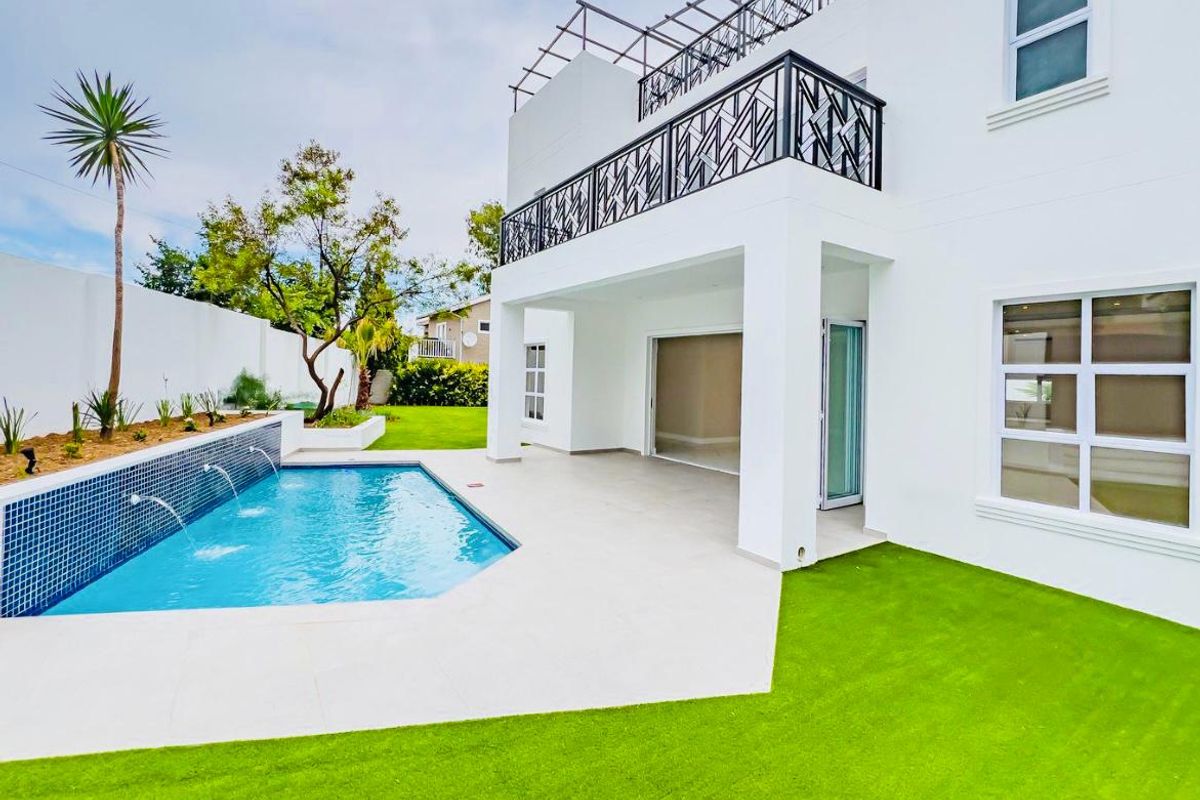 A modern rear exterior featuring a swimming pool with water features, a covered patio, and a balcony with decorative metal railings.