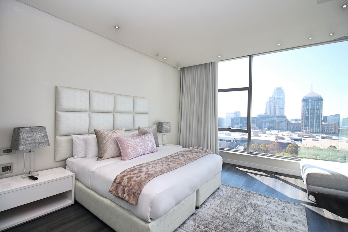 A modern, well-lit bedroom featuring floor-to-ceiling windows with expansive city views, recessed ceiling lighting, and high-quality dark wood flooring.