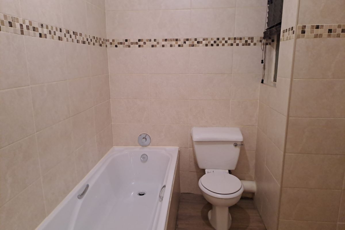 A functional bathroom featuring a built-in bathtub and a standard toilet, finished with neutral wall tiles and a decorative mosaic border.