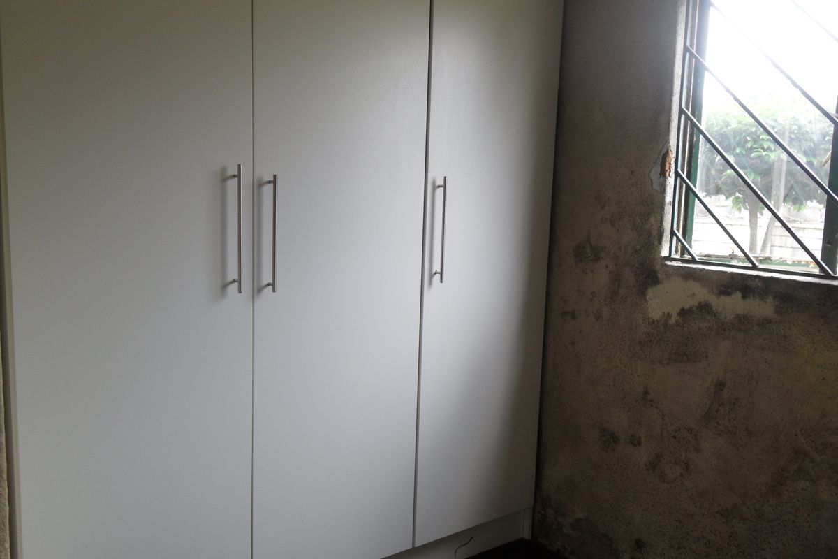 A bedroom featuring built-in white cabinetry, showing significant wall surface damage and damp staining around the window area requiring repair.