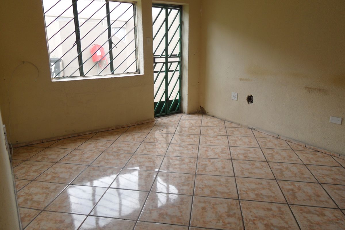 An empty living room featuring tiled flooring, security bars on the window and door, and visible wall damage requiring repair.