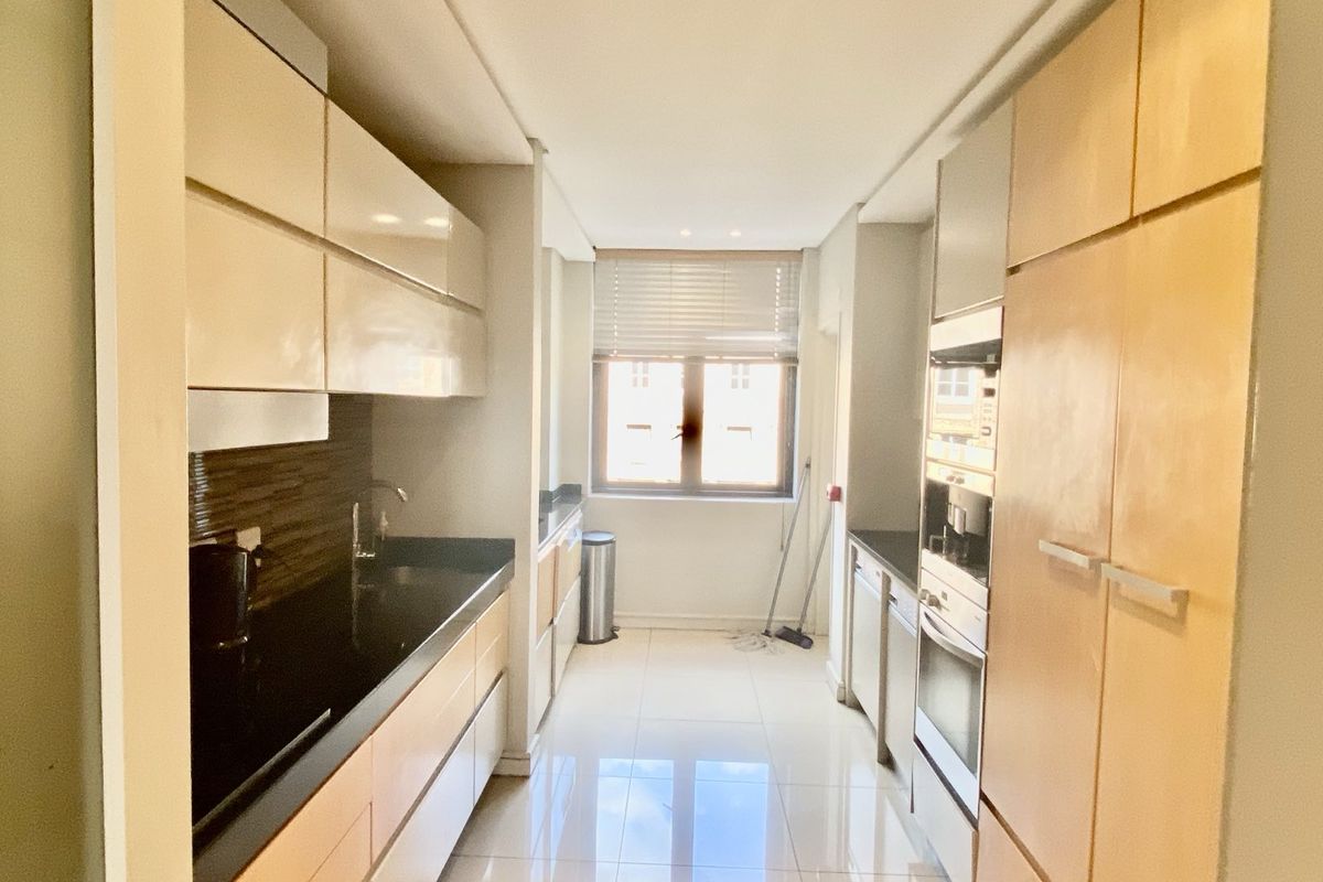 A galley-style kitchen featuring light wood cabinetry, dark stone countertops, integrated oven, and tiled flooring with natural light from a window.