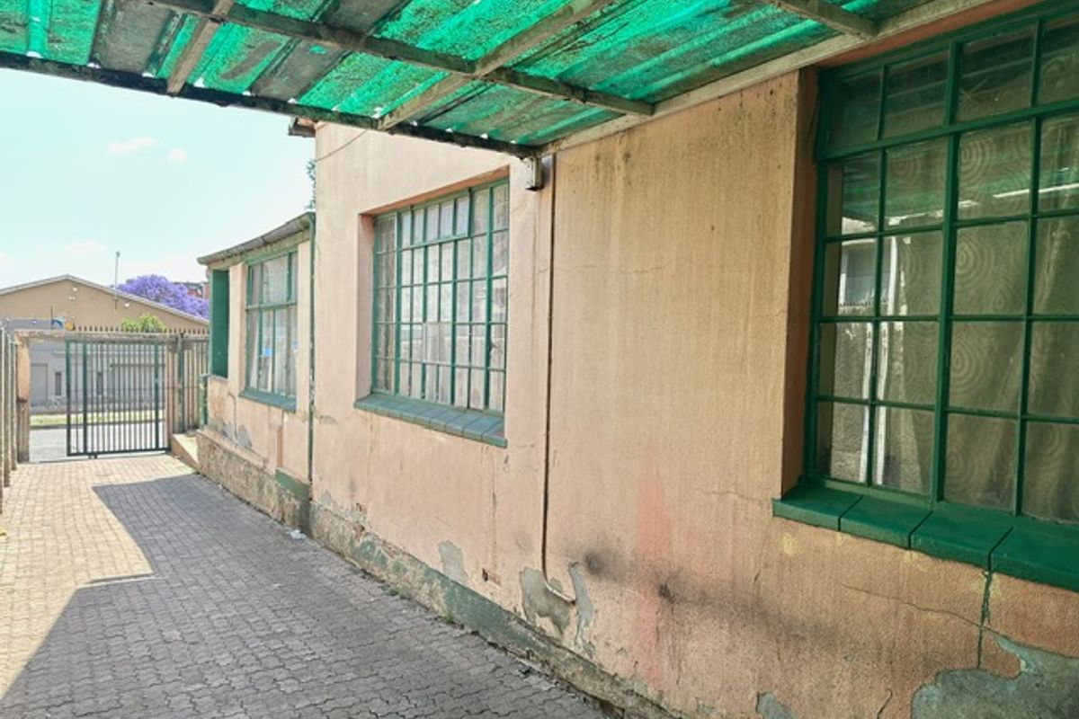 Side exterior view of the property featuring a paved walkway, green metal-framed windows, and a covered outdoor area with a corrugated plastic roof.