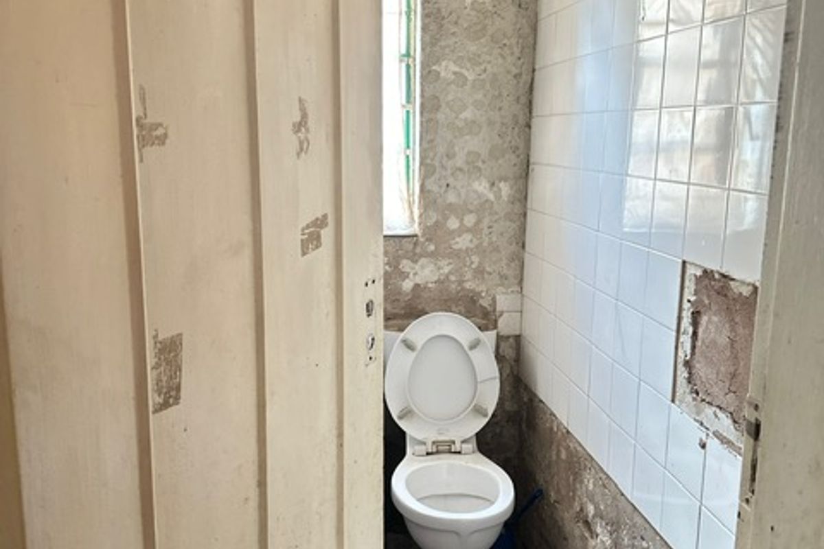 A small, dated bathroom requiring significant renovation, featuring a standard toilet and partially tiled walls with visible surface damage.