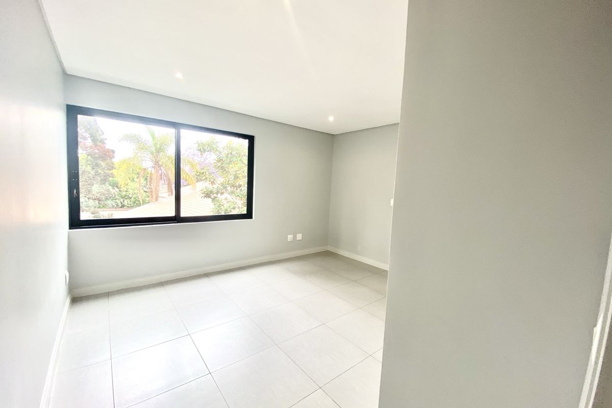 A bright, empty bedroom featuring neutral tiled flooring, recessed ceiling lighting, and a large window with a black frame overlooking the garden.