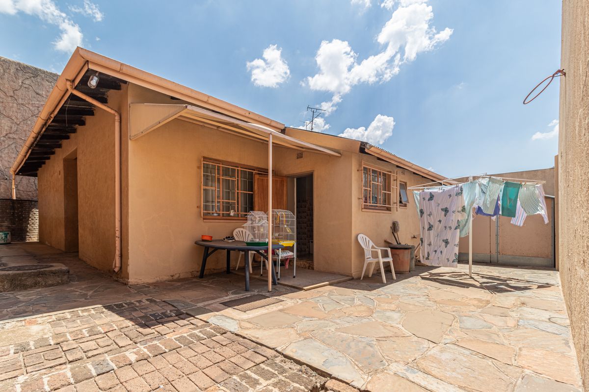 A rear exterior view of the property featuring a paved courtyard area, metal security window bars, and a covered patio section.