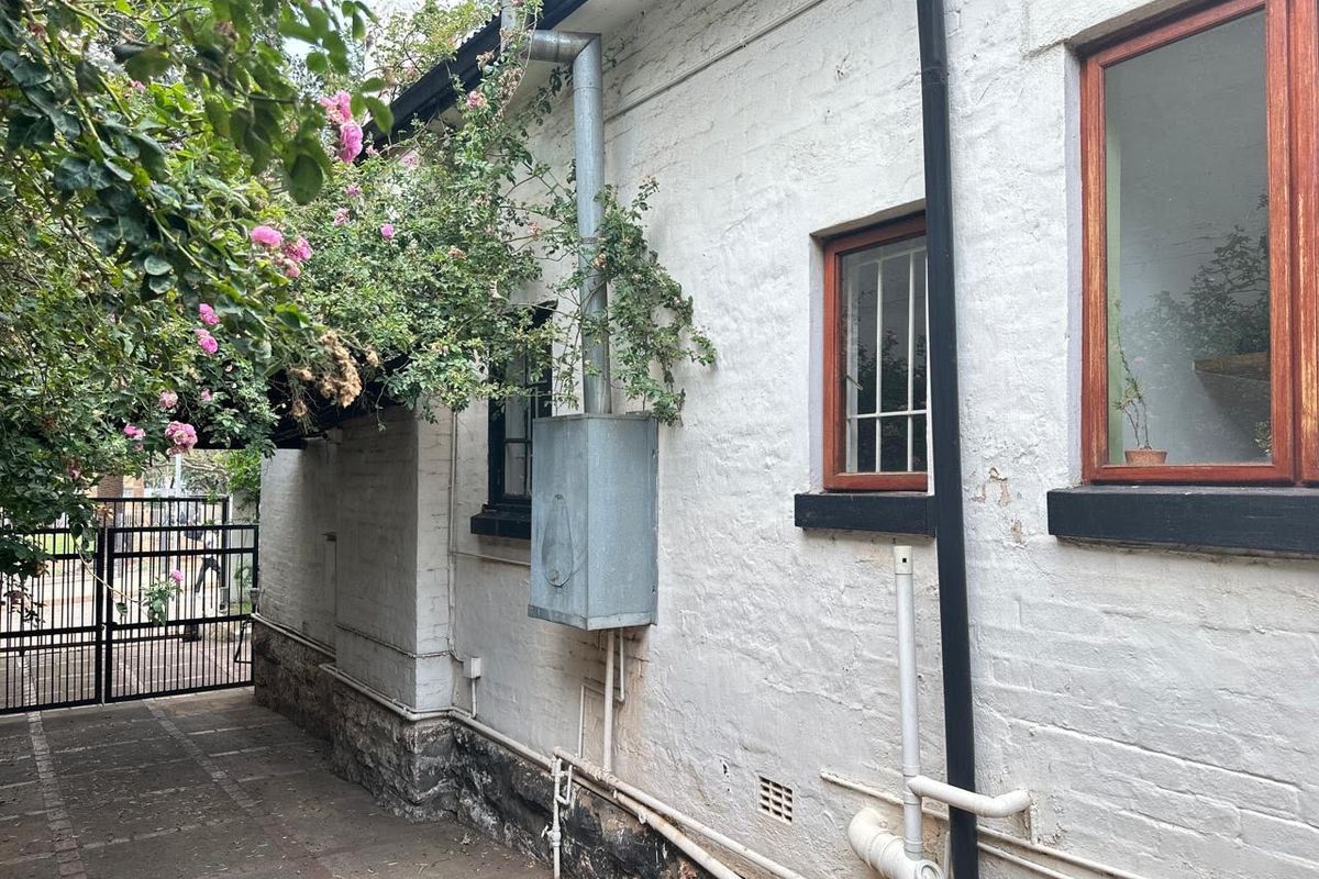 Side exterior view of the property featuring painted brick walls, timber-framed windows with security bars, and exposed utility piping.