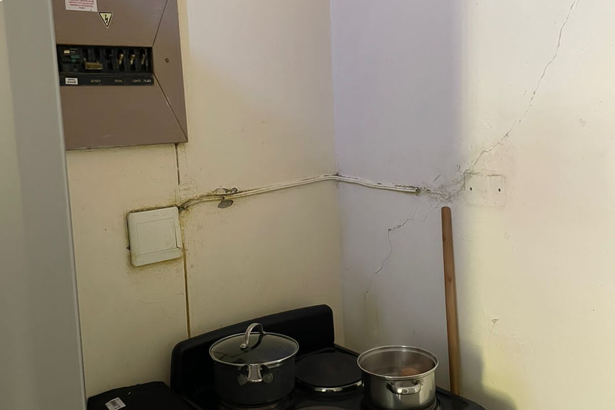 A small kitchen corner featuring a freestanding electric stove, wall-mounted electrical distribution board, and visible structural wall cracking.
