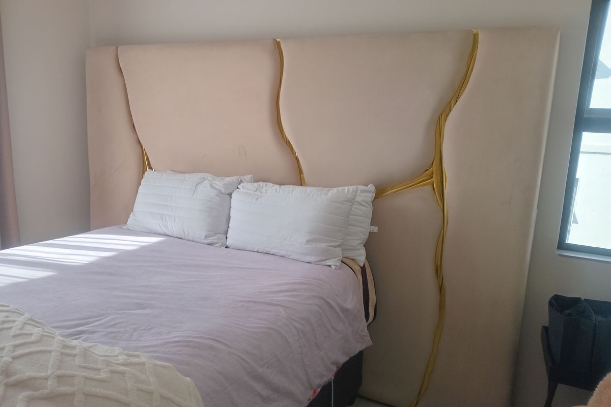 A bedroom featuring a large, custom-designed upholstered headboard with gold accent detailing, positioned against a neutral wall with natural light from a side window.