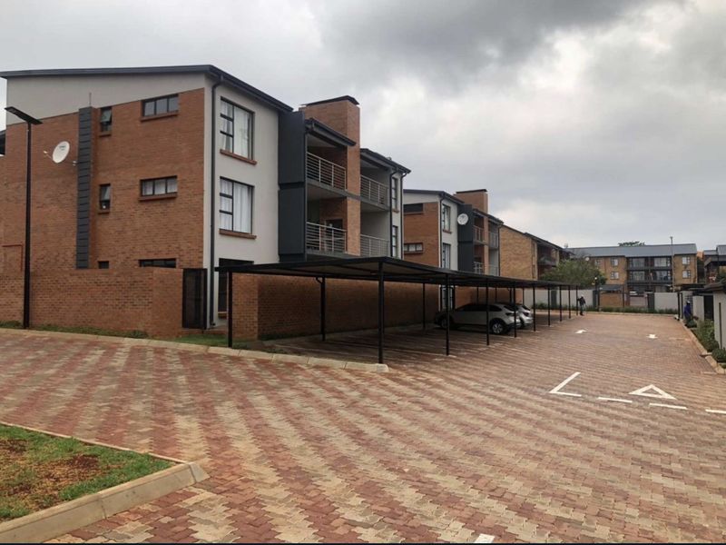 Exterior view of a modern apartment complex featuring brick facade buildings, private balconies, and dedicated covered parking bays on a paved driveway.