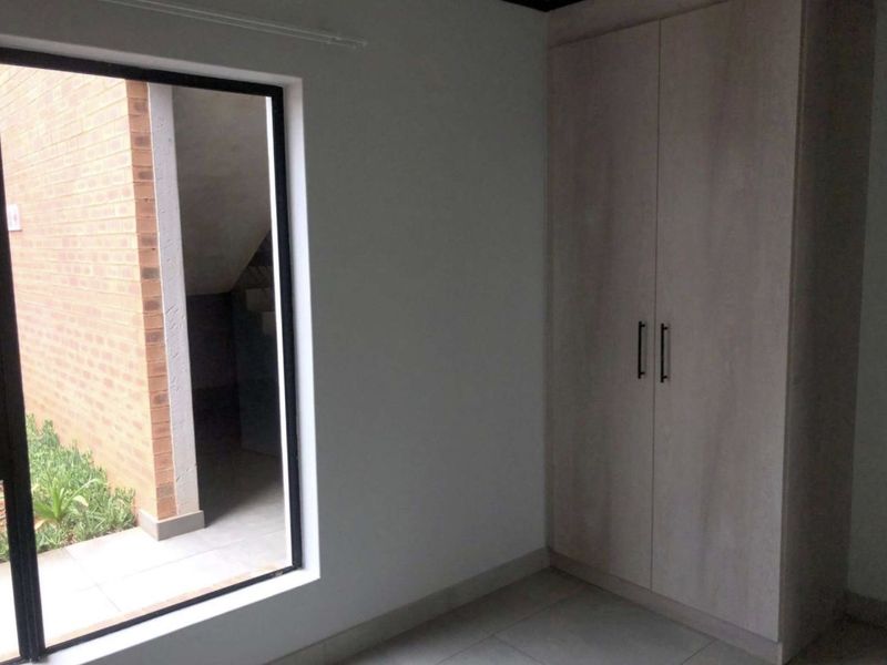 A clean, empty bedroom featuring neutral tiled flooring, a built-in wardrobe, and a large window providing natural light and a view of the exterior.