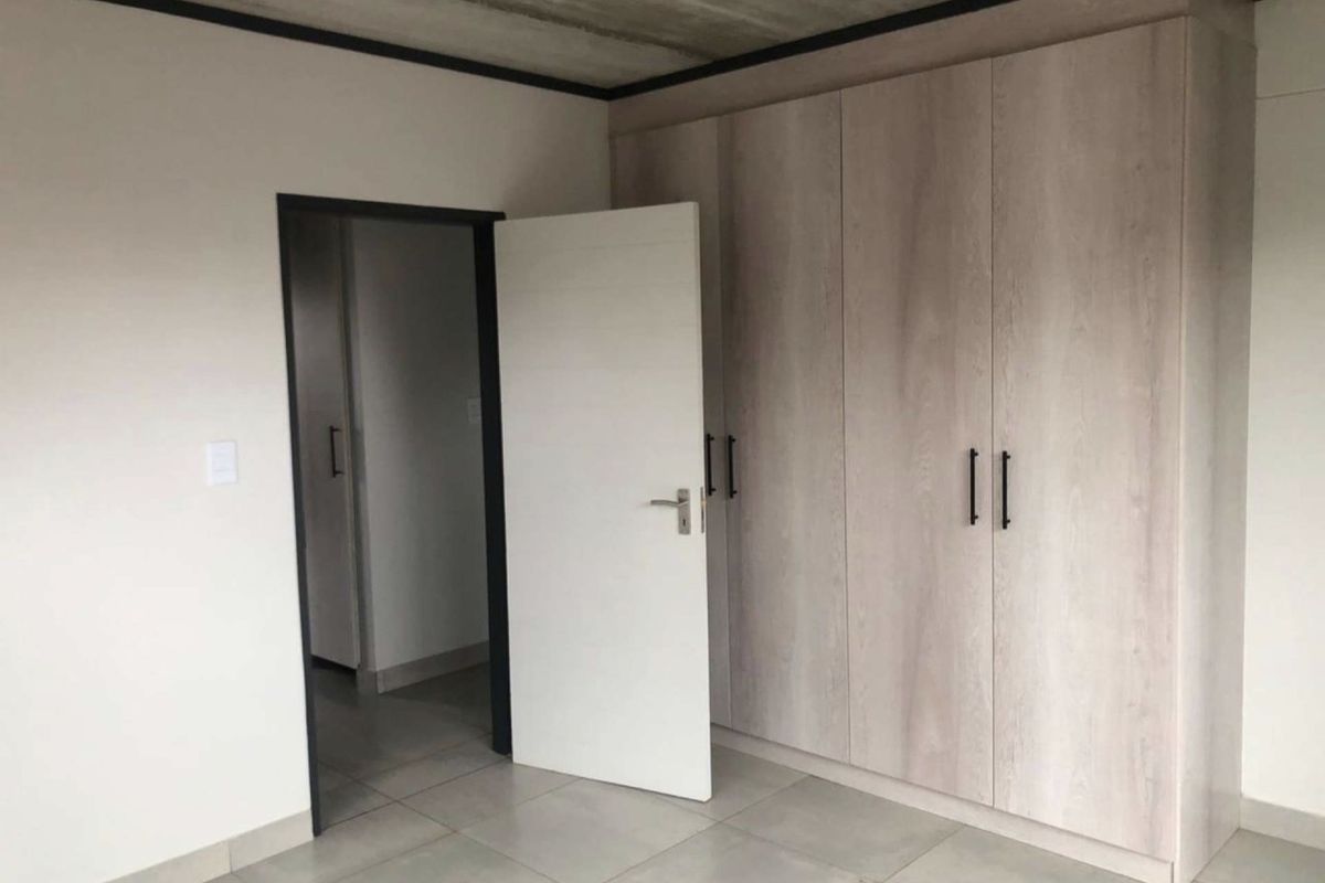 A clean, modern bedroom featuring built-in light wood-grain cupboards, neutral floor tiles, and an industrial-style exposed concrete ceiling.