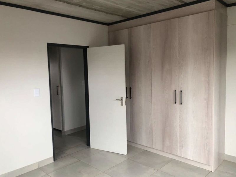 A clean, modern bedroom featuring built-in light wood-grain cupboards, neutral floor tiles, and an industrial-style exposed concrete ceiling.