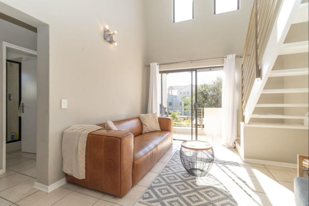 Bright living area featuring double-volume ceilings, a staircase with metal balustrades, tiled flooring, and sliding glass doors leading to a balcony.