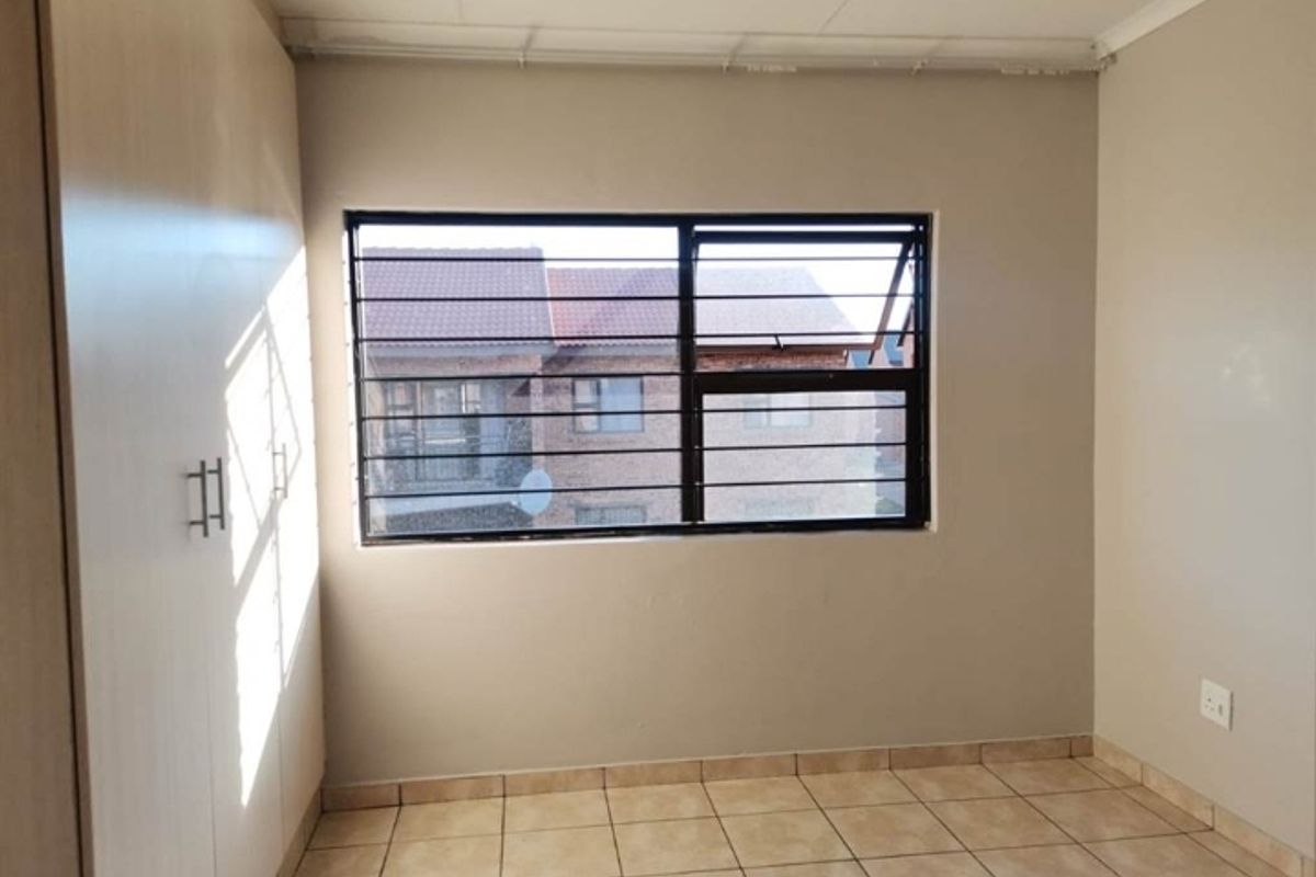 A bright, empty bedroom featuring built-in cupboards, tiled flooring, and a window fitted with security bars.