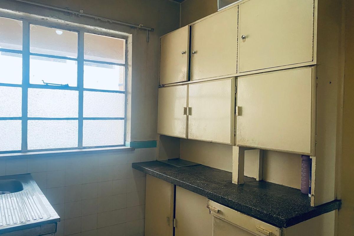 A dated kitchen area featuring original cabinetry, a laminate countertop, and a stainless steel sink unit with a large window providing natural light.